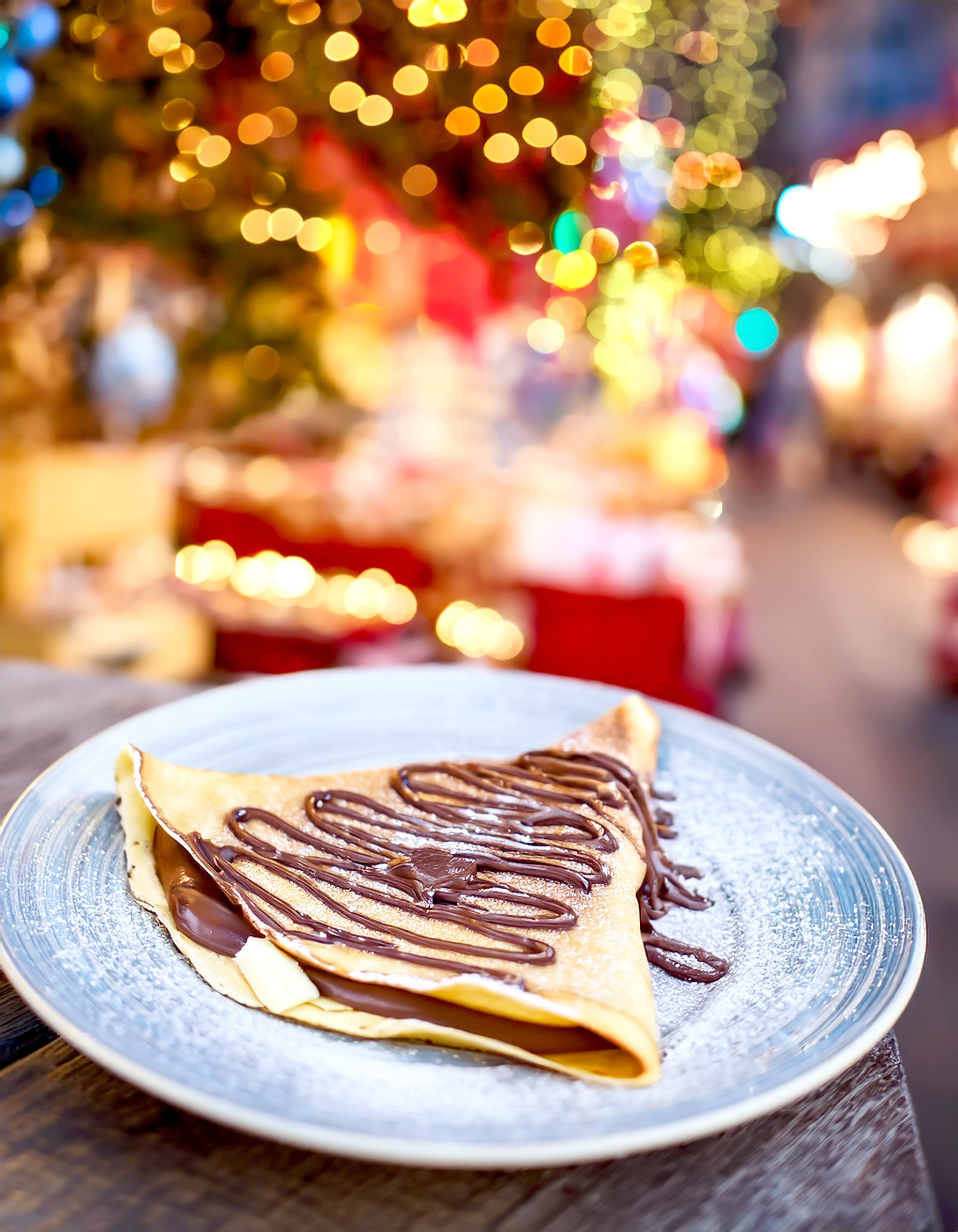 Ein Crepes mit Schokosoße auf einem weißen Teller, im Hintergrund weihnachtliche Beleuchtung